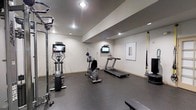 This fitness center offers a dedicated space for a comprehensive workout experience within a clean and modern environment. The room features durable rubber flooring designed to withstand heavy equipment and high-impact activity, providing both safety and comfort underfoot. Recessed lighting is installed throughout the ceiling, ensuring the space is well-lit for all types of exercise.

The cardio section includes a treadmill and an upright stationary bike, each positioned for easy use and equipped with individual wall-mounted flat-screen televisions, allowing users to stay entertained during their workout. A multi-function cable machine and a dedicated elliptical trainer round out the strength and low-impact cardio options available.

Suspension training straps are anchored to the wall, providing a versatile tool for bodyweight-based strength and stability exercises. A weight bench is also present, supporting a variety of strength training routines. Framed artwork on the wall adds a subtle aesthetic touch, softening the utilitarian nature of the space.

The neutral color palette of white walls and a gray ceiling contributes to an open, focused atmosphere. Overall, this fitness center is thoughtfully arranged to accommodate a wide range of fitness activities in an organized and functional layout.