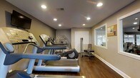 Located at 1660 Old Trolley Rd in Summerville, South Carolina, this fitness center offers a well-equipped and thoughtfully designed exercise space on a single floor. The room features rich hardwood flooring that lends warmth and durability underfoot, complemented by neutral-toned walls that create a clean, focused environment for working out. Recessed lighting is installed throughout the ceiling, providing bright, even illumination across the entire space. The fitness area is outfitted with a selection of cardio machines, including treadmills and an elliptical trainer, arranged to allow comfortable circulation around each piece of equipment. Strength training is also accommodated with a dedicated weight and resistance machine, as well as an adjustable bench, making the space suitable for a well-rounded workout routine. A large interior window on one wall connects the room visually to an adjacent area, adding openness to the space. A white paneled door provides access, contributing to the room's clean and organized aesthetic. The overall ambiance is practical yet inviting, designed to support an active lifestyle with all essential amenities in a compact and efficient layout.