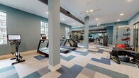 Located at 806 16th Ave N in Nashville, TN, this fitness center offers a generously sized, single-floor space designed to support a full range of workout routines. The open floor plan is finished with a distinctive patchwork carpet tile pattern in shades of blue, gray, and cream, lending the space a modern, polished aesthetic. Walls painted in a calming teal-blue tone complement the bright, recessed lighting and ceiling fans that maintain a comfortable environment throughout.

Large grid-pane windows line the perimeter, flooding the space with natural light and providing views of the surrounding outdoor area. The cardio zone features multiple treadmills with integrated display screens, elliptical machines, a rowing machine, and a stationary bike, all positioned to take advantage of the natural light. A dedicated free weights area houses a full rack of dumbbells alongside a flat bench, with floor-to-ceiling mirrors and a wall-mounted flat-screen TV creating an expansive, motivating atmosphere.

The strength training section is equally well-appointed, with a multi-functional Smith machine, cable systems, and a variety of resistance equipment. Additional functional fitness tools, including medicine balls, kettlebells, and stability equipment, round out the facility's comprehensive offerings. Exposed ductwork and structural columns add an industrial character that balances the space's clean, contemporary finishes.