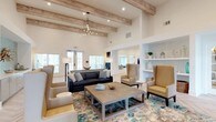 Parkside At The Beach's clubhouse is a thoughtfully designed single-story gathering space that blends coastal charm with modern functionality. The main lounge commands attention with soaring ceilings accented by rustic wood beams and an elegant chandelier, creating a grand yet welcoming atmosphere. Large windows flood the space with natural light, while a navy sofa, neutral wingback chairs, and a concrete coffee table atop a blue-and-ivory area rug establish a sophisticated coastal aesthetic.

Adjacent areas transition seamlessly through arched openings and French doors, revealing a private meeting nook finished in soft seafoam tones. A round glass-top table with a sculptural driftwood base sits beneath a striking geometric pendant light, complemented by a sleek credenza. Herringbone-patterned light wood flooring flows throughout, unifying the space.

A dedicated lounge and coworking area features built-in bar-height workstations, a wall-mounted television, and comfortable swivel chairs arranged around a warm wood accent table. A separate media lounge with seafoam accent walls, sconce lighting, and additional seating offers an ideal setting for casual gatherings. The kitchen area is appointed with light wood cabinetry, quartz countertops, a stainless steel double sink, and a sea-glass mosaic tile backsplash, completing the refined beachside ambiance.