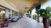 This covered outdoor terrace is designed for comfortable communal gathering in a tropical setting. The space features a generously sized, tile-floored lanai that runs the length of the building, offering an open yet sheltered environment ideal for relaxation and socializing. Overhead, multiple ceiling fans with decorative wooden blades provide consistent airflow, keeping the area comfortable year-round. Recessed lighting supplements natural light throughout the covered section.

The lounge area is furnished with wicker-style seating upholstered in navy blue and patterned fabric, accompanied by rich mahogany-toned wooden coffee tables. Blue decorative outdoor drapery panels frame the arched openings along the perimeter, adding a resort-like elegance while offering optional shade and privacy. Large, tall planters filled with bold tropical foliage—including fan palms and elephant ear plants—are strategically placed to enhance the lush, verdant ambiance.

A wrought iron railing lines the open sides of the terrace, beyond which mature palm trees and well-manicured landscaping are visible, reinforcing the tropical character of the setting. A series of blue-framed glass French doors connect the terrace to an interior space, allowing for seamless indoor-outdoor flow. Overall, the terrace delivers a refined, resort-inspired atmosphere suited for both quiet retreat and social gatherings.