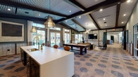 This single-floor community clubhouse delivers a sophisticated blend of industrial and modern design throughout its open, well-appointed layout. A distinctive hexagonal tile floor in warm earth tones runs continuously across the space, anchoring each zone with a cohesive aesthetic. Exposed dark steel beams and corrugated metal ceiling panels add industrial character, while recessed lighting, statement pendant fixtures, and a sculptural sputnik chandelier provide layered illumination throughout.

The entry foyer welcomes visitors with soaring ceilings, floor-to-ceiling glazed doors that invite abundant natural light, and an elegant beaded chandelier. Adjacent, a fully equipped kitchen serves as a functional centerpiece, featuring rich walnut cabinetry, dark subway tile backsplash, stainless steel appliances, and a large white-topped island with built-in seating for casual gathering. Open shelving displays curated décor alongside practical storage.

The main lounge area offers versatile social spaces, including a full-size billiards table positioned beneath industrial pendant lights and a comfortable seating arrangement with a wall-mounted television. Textured, panel-clad walls and large windows framing outdoor views enhance the lounge's inviting ambiance. A teal barn door adds a bold accent, leading to additional areas within the facility. Together, these spaces create an engaging, design-forward environment suited for both relaxation and socializing.
