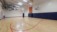 This indoor basketball court is a purpose-built athletic facility designed to support both competitive play and recreational activity. The centerpiece of the space is a polished hardwood maple floor, finished with clearly marked court lines in red and orange, including a painted perimeter boundary stripe and a branded center-court logo. The generous floor area provides ample room for full-court play and drills. Safety is a prominent design consideration throughout: thick navy blue padded wall mats line the perimeter walls, offering protection for players near the boundaries, while padded column covers safeguard structural posts on the court. Regulation glass-backboard basketball hoops are mounted at each end of the court, and a rack of basketballs is conveniently stored along the sideline for immediate use. The ceiling is elevated, providing the vertical clearance required for unobstructed gameplay, and features exposed ductwork, industrial-grade ventilation systems, and fluorescent lighting fixtures that ensure bright, even illumination across the entire court surface. Accent walls in a bold terracotta orange add a dynamic visual contrast to the crisp white walls and navy padding. The overall environment is clean, functional, and well-maintained, making it a highly capable facility for basketball, fitness training, and multi-sport use.