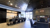 This single-floor space makes a strong architectural statement through its dramatically vaulted ceiling clad in dark wood paneling, which adds warmth and visual depth while amplifying the sense of height throughout the interior. Pendant lighting hangs at varying lengths from the ceiling, casting focused illumination across the space and complementing the recessed lighting integrated throughout.

The layout is thoughtfully organized into distinct functional zones. A bar and kitchenette area along one wall features a stone backsplash, open shelving, and stainless steel appliances, with bar-height seating providing an inviting spot for casual gathering. Adjacent dining tables and chairs offer additional seating for groups.

Toward the far end of the room, a sleek linear fireplace is set against a stone accent wall, flanked by upholstered lounge chairs that create a relaxed seating area. Large windows and glass doors nearby allow natural light to filter in, brightening the space and providing a visual connection to the outdoors.

The flooring throughout features light-toned wood planks that contrast effectively with the darker ceiling and accent walls. The overall ambiance is contemporary and refined, balancing industrial and residential design elements cohesively.