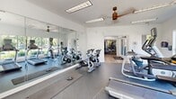 Located at 21222 Nautique Blvd in Cornelius, NC, this fitness center offers a comprehensive workout facility spread across a single level. The space is organized into two distinct exercise rooms connected by a welcoming entrance foyer, which features warm hardwood-style flooring, decorative columns, soaring ceilings, and a water fountain alcove with stylish mosaic tile detail.

The first exercise room is dedicated to strength training, housing a multi-station cable machine, free weights, benches, and a variety of resistance equipment. Rubber-textured flooring provides safety and durability throughout, while ceiling fans and recessed panel lighting ensure a comfortable environment. Large windows allow natural light to fill the room, and wall-mounted mirrors amplify the sense of space.

The second exercise room focuses on cardio and cycling, featuring multiple treadmills, elliptical machines, and spin bikes arranged for easy access. Wall-mounted televisions keep members entertained during workouts, and full-length mirrors along one wall further enhance the open feel. Natural light streams in through generously sized windows, creating a bright and energizing atmosphere.

Together, the two rooms deliver a well-rounded fitness experience with a clean, modern aesthetic suited for residents of the surrounding community.