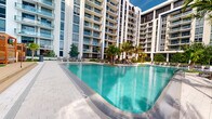 This contemporary residential complex is defined by its striking modern architecture, characterized by clean white facades, floor-to-ceiling glazing, and private balconies that span across multiple high-rise towers. The buildings are arranged in a thoughtful configuration that frames a central outdoor amenity area, creating a semi-enclosed courtyard environment.

The centerpiece of the complex is a generously proportioned resort-style swimming pool with vivid turquoise water, finished with a sleek tiled surimeter and smooth paved surrounds. The pool deck is furnished with lounge chairs and sun loungers, offering ample space for relaxation. Tall, mature palm trees are strategically planted throughout the courtyard, lending a tropical ambiance and providing natural shade across the deck area.

The surrounding towers rise prominently, each featuring uniform rows of glass-railed balconies that overlook the pool below. The exterior design employs a consistent palette of white and light grey tones accented by dark window framing and architectural fins, contributing to a polished, upscale aesthetic. The open-air setting allows for abundant natural light and open sky views, enhancing the resort-like character of the shared outdoor space.