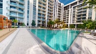 This contemporary residential complex is defined by its striking modern architecture, characterized by clean white facades, floor-to-ceiling glazing, and private balconies that span across multiple high-rise towers. The buildings are arranged in a thoughtful configuration that frames a central outdoor amenity area, creating a semi-enclosed courtyard environment.

The centerpiece of the complex is a generously proportioned resort-style swimming pool with vivid turquoise water, finished with a sleek tiled surimeter and smooth paved surrounds. The pool deck is furnished with lounge chairs and sun loungers, offering ample space for relaxation. Tall, mature palm trees are strategically planted throughout the courtyard, lending a tropical ambiance and providing natural shade across the deck area.

The surrounding towers rise prominently, each featuring uniform rows of glass-railed balconies that overlook the pool below. The exterior design employs a consistent palette of white and light grey tones accented by dark window framing and architectural fins, contributing to a polished, upscale aesthetic. The open-air setting allows for abundant natural light and open sky views, enhancing the resort-like character of the shared outdoor space.