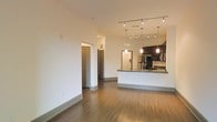 This single-floor unit showcases an open-concept design that integrates the living and kitchen areas into one cohesive, flowing space. Warm hardwood flooring extends throughout, lending a clean and contemporary aesthetic to the interior.

The kitchen serves as a focal point, featuring dark espresso cabinetry that contrasts sharply against the white walls and light countertops. A peninsula-style breakfast bar with an integrated sink provides both a functional workspace and a casual dining option. Overhead, a combination of track lighting and elegant pendant lights illuminates the kitchen, creating a layered and well-lit environment. Stainless steel appliances are visible, adding a modern, polished touch.

The adjoining living area is generously proportioned, bathed in ambient light, and benefits from the open sightlines created by the layout. Crisp white walls and high baseboards contribute to a bright, airy atmosphere throughout.

A hallway visible from the main living space leads to additional rooms, hinting at a thoughtful and functional floor plan. The overall ambiance is modern and inviting, with clean lines and quality finishes throughout.