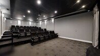 Located at 1061 Spring Creek Dr in Forest, VA, this dedicated home theater space delivers an authentic cinema experience within a single, thoughtfully designed room. The space is configured with multiple rows of plush, dark leather recliner-style seats arranged in a tiered layout, ensuring clear sightlines to the large projection screen from every position. The screen is elegantly framed by floor-length silver-gray drape curtains, adding a classic theatrical touch to the room's aesthetic. A ceiling-mounted projector anchors the center of the dark, matte black ceiling, which is trimmed with recessed accent lighting and wall-mounted sconces that cast a soft, ambient glow along the gray walls. The contrast between the dark ceiling and lighter walls creates a dramatic, immersive atmosphere well-suited for screenings. Patterned carpet with a subtle geometric design covers the floor throughout, contributing to both the visual appeal and acoustic quality of the room. A raised platform separates the rear seating rows from the front, enhancing the stadium-style viewing arrangement. The overall design is polished and functional, purpose-built to replicate the feel of a commercial movie theater in a private setting.