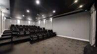 Located at 1061 Spring Creek Dr in Forest, VA, this dedicated home theater space delivers an authentic cinema experience within a single, thoughtfully designed room. The space is configured with multiple rows of plush, dark leather recliner-style seats arranged in a tiered layout, ensuring clear sightlines to the large projection screen from every position. The screen is elegantly framed by floor-length silver-gray drape curtains, adding a classic theatrical touch to the room's aesthetic. A ceiling-mounted projector anchors the center of the dark, matte black ceiling, which is trimmed with recessed accent lighting and wall-mounted sconces that cast a soft, ambient glow along the gray walls. The contrast between the dark ceiling and lighter walls creates a dramatic, immersive atmosphere well-suited for screenings. Patterned carpet with a subtle geometric design covers the floor throughout, contributing to both the visual appeal and acoustic quality of the room. A raised platform separates the rear seating rows from the front, enhancing the stadium-style viewing arrangement. The overall design is polished and functional, purpose-built to replicate the feel of a commercial movie theater in a private setting.