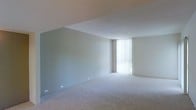 This single-floor space presents a clean, minimalist aesthetic defined by neutral color tones and straightforward design elements. The main living area is notably open and spacious, featuring wall-to-wall carpeting in a warm beige tone that extends throughout the space, providing a soft and cohesive feel underfoot. The walls are finished in a muted sage-gray hue, lending a calm and understated ambiance to the interior.

A standout feature of the main room is the expansive floor-to-ceiling window dressed with vertical blinds, which allows an abundance of natural light to filter into the space, creating a bright and airy atmosphere. The smooth, textured ceiling complements the clean wall finishes throughout.

A hallway connects to additional areas of the space and showcases a built-in shelving unit with multiple open shelves, offering practical storage and display opportunities. The hallway leads toward the main living area, with the large window visible in the background, drawing the eye forward and reinforcing the sense of depth and openness. Overall, the space offers a neutral, move-in-ready canvas well-suited for a variety of interior styles.