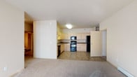 This single-floor residence offers a practical and comfortable open-concept layout that seamlessly connects the kitchen and living areas. The kitchen is well-appointed with light maple cabinetry that extends to the upper walls, providing generous storage space. Dark granite-style countertops offer a contrasting accent, while the suite of black appliances — including a refrigerator, electric range, built-in microwave, and dishwasher — delivers full functionality. The tile flooring in the kitchen is durable and easy to maintain, transitioning smoothly into the plush wall-to-wall carpeting of the adjoining living area. The living space is open and airy, benefiting from a neutral color palette of soft whites and warm creams that lend a clean, inviting atmosphere. Flush-mounted ceiling lighting provides consistent illumination throughout. A partially enclosed wall creates a subtle division between the kitchen and living zones while preserving the open flow of the floor plan. A doorway visible to the right suggests access to additional rooms, rounding out this well-organized, move-in-ready living space designed for everyday comfort.