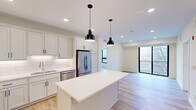 This thoughtfully designed 2-bedroom, 2-bathroom residence features a clean, contemporary aesthetic throughout its single-floor layout. The open-concept kitchen serves as the heart of the home, outfitted with white shaker cabinetry, white quartz countertops, a subway tile backsplash, stainless steel appliances including a French door refrigerator, and a central island with pendant lighting overhead. Light hardwood flooring flows through the main living areas, complementing the bright, neutral palette. The living area extends toward a glass door that opens to a private balcony, filling the space with natural light and outdoor views of surrounding trees. The primary bedroom features carpeted flooring, a large window framing treetop views, and direct access to a well-appointed en suite bathroom with a dual-sink vanity, black hardware accents, a separate walk-in shower, and a dedicated closet. The second bedroom is equally bright with a large window and plush carpeting. The second bathroom offers a single vanity and a full tub-shower combo. A dedicated in-unit laundry room with a full-size washer and dryer adds everyday convenience. Two hallways provide thoughtful circulation throughout the home, completing this practical and modern residence.