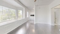 Located at 1501 W Adams Ave in Los Angeles, CA, this single-floor unit offers a well-organized layout comprising a living room, bathroom, closet, and hallway. The living room is a standout space, featuring gleaming gray hardwood floors, crisp white walls, and a textured ceiling with a ceiling fan. Multiple windows flood the room with natural light and frame views of the surrounding greenery, creating a bright and airy atmosphere. Wainscoting panels beneath the windows add a classic architectural detail, while a wall-mounted heater provides functional comfort. The space transitions seamlessly toward a compact kitchen area, visible from the living room, which features white cabinetry and a bold dark tile backsplash — a striking design contrast. A secondary room with hardwood flooring and a window offers additional flexibility as a sleeping or office space. The bathroom is finished with elegant marble-style porcelain tile flooring in white and gray tones, white subway-tiled shower walls, and clean white fixtures, delivering a polished, cohesive aesthetic. The hallway connects the spaces efficiently, contributing to the unit's practical and straightforward flow. Overall, the interior presents a clean, bright, and modern sensibility throughout.