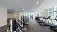 This contemporary fitness center is thoughtfully designed to support a complete workout experience within a single, open-plan level. The space is defined by its expansive floor-to-ceiling windows that line an entire wall, flooding the interior with natural light and offering views of the surrounding building complex and landscaping. The clean-lined architectural design features smooth white walls, recessed lighting, and durable rubber flooring throughout the exercise areas, creating a polished yet functional environment.

The cardio zone is well-appointed with a row of True-brand treadmills, elliptical trainers, stationary bikes, and a recumbent cycle, all positioned directly in front of the large windows to maximize natural light during workouts. The strength training area features multiple cable and resistance machines, a free weight section with a full dumbbell rack, adjustable benches, and open floor space for functional training.

Adjacent to the main fitness floor is a stylish lounge area with a large white island countertop and bar-style seating, set against bright walls and additional window access. This area provides a relaxed space for pre- or post-workout use. The overall ambiance is bright, modern, and clean, reflecting a well-maintained, professionally outfitted facility.