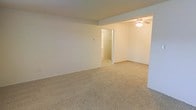 This single-floor space presents a clean, straightforward layout finished in a neutral palette throughout. The main living area is carpeted in a warm beige tone that extends across the generous open floor plan, creating a cohesive and understated foundation. White walls provide a bright, versatile backdrop well-suited for a variety of furnishing arrangements.

A ceiling fan with an integrated light fixture is installed in one section of the space, offering both overhead illumination and air circulation. A subtle change in ceiling height or a structural beam subtly divides the room into distinct functional zones while maintaining an open feel between them.

A doorway connects to an adjacent room, visible from the main area and similarly finished with matching flooring and light-toned walls. This connected space appears to function as a secondary room or hallway, with what appears to be a closet accessible from it, adding practical storage to the layout.

Overall, the space conveys a simple, move-in-ready character with consistent finishes throughout, offering a neutral and functional environment suitable for a range of uses.