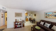 This single-floor residence offers a functional and comfortable layout connecting several distinct living areas. The open-concept living and dining space forms the heart of the home, featuring carpet flooring, a textured ceiling, and neutral walls that lend a warm, understated ambiance. The living area accommodates a full sofa and coffee table, while a pendant light anchors the adjacent dining space, which transitions to tile flooring and offers room for a multi-seat dining set. Framed artwork decorates the walls throughout, adding character to the space.

The kitchen is compact yet practical, featuring warm wood cabinetry extending to the upper walls for generous storage, a white drop-in sink, white appliances including a refrigerator and range, and light countertops. A secondary room near the entry serves as a flexible space, currently used as a sitting or hobby room, and includes a closet, exposed brick-style walls, carpeted floors, and a window with natural light.

The bedroom is well-proportioned with brick-style accent walls, carpet, a window with blinds, and a ceiling light fixture. The full bathroom includes a white vanity with a mirrored medicine cabinet, a tub-shower combination with a curtain, and tile flooring. The entry area features a large window, providing a bright welcome into the unit.