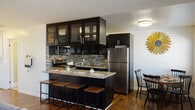 This well-designed single-floor residence offers a cohesive blend of modern style and comfortable living throughout its thoughtfully arranged spaces. The kitchen is a standout feature, showcasing dark espresso cabinetry with glass-front uppers, a mosaic tile backsplash, granite countertops, and a full suite of stainless steel appliances including a gas range, built-in microwave, and refrigerator. A breakfast bar with wood-topped stools provides casual seating and connects seamlessly to the dining area, which features a sunny window with blinds and decorative accents. Rich hardwood flooring flows throughout the main living areas, grounding the space with warmth and continuity. The living room is finished in a sophisticated slate-blue tone, complemented by a ceiling fan, wall-mounted television, and tasteful décor. The bedrooms continue the blue-gray color palette, each fitted with ceiling fans, hardwood floors, and windows that invite natural light. The primary bedroom is generously proportioned, offering ample space for a full furniture arrangement. The bathroom is finished with large-format stone-look tile surrounding a soaking tub and shower combination enclosed by sliding glass doors, delivering a clean, spa-inspired aesthetic throughout.