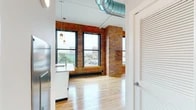 Located at 1 Websters Landing in downtown Syracuse, NY, this urban loft-style unit masterfully blends industrial character with modern finishes across a single thoughtfully laid-out floor. Soaring ceilings, exposed ductwork, and original brick columns and accent walls pay homage to the building's industrial heritage, while light hardwood flooring and crisp white walls create a clean, contemporary contrast throughout.

The open-concept kitchen and living area serves as the heart of the space, anchored by a large center island with striking granite countertops and an undermount sink. White shaker cabinetry pairs seamlessly with stainless steel appliances, including a full-size refrigerator, electric range, and built-in microwave. Oversized black-framed windows flood the space with natural light and frame sweeping views of the Syracuse skyline and surrounding cityscape. A stylish barn door adds a refined industrial touch to the living area.

The bathroom features granite countertops, white cabinetry, a large mirror, and tile flooring. Two closets and a hallway provide practical storage solutions. Pendant lighting throughout adds warmth and character to this distinctive urban retreat.