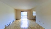 This single-floor space showcases a clean, functional design characterized by an open layout and abundant natural light. The main living area is anchored by polished hardwood floors that reflect sunlight, creating a warm and inviting atmosphere throughout. A large sliding glass door dominates one wall, providing direct access to an outdoor balcony while framing views of lush greenery beyond. Baseboard heating units run along the perimeter walls, ensuring efficient climate control throughout the colder months. The walls are finished in a neutral off-white tone, offering a versatile backdrop adaptable to a variety of interior styles. An open doorway connects the living area to an adjacent room, which features its own window that brings in additional natural light and contributes to the overall sense of airiness. The layout flows naturally between spaces, supporting both everyday living and easy movement throughout. The combination of hardwood flooring, generous glazing, and a straightforward architectural design gives the space a timeless, practical character suited to a range of uses.