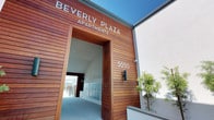 Beverly Plaza Apartments presents a striking first impression through its contemporary architectural design. The entrance facade is clad in warm, rich horizontal wood planking that frames a generously proportioned entryway, creating a bold yet welcoming statement. The building number 5050 is cleanly mounted against the contrasting white stucco exterior, reinforcing the modern aesthetic.

The entry corridor is bright and airy, benefiting from an open-air design that allows natural light to flow through freely. A bank of individual mailboxes lines one wall of the lobby area, offering practical convenience for residents. The flooring throughout the entry is finished in light-toned large-format tile, contributing to a clean, polished appearance.

Beyond the entry, a second opening frames views of the surrounding neighborhood, including mature palm trees and neighboring low-rise structures, lending a sense of openness and connection to the outdoors. Sleek metal railings border a stepped pathway leading outward, flanked by landscaped planters with ornamental grasses and greenery. The overall ambiance is modern, well-maintained, and thoughtfully designed, balancing natural materials with contemporary finishes.