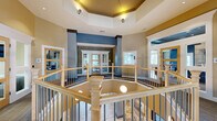 Located at Ivystone Square in Chesapeake, Virginia, this impressive two-story community clubhouse offers a thoughtfully designed gathering space that combines elegance with functionality. The main floor commands attention with its sweeping open layout, featuring rich herringbone-patterned hardwood flooring, tray ceilings with recessed lighting, and a bold color palette of deep blues, warm golds, and crisp whites that lend the space a polished, contemporary feel. A welcoming lounge area is anchored by large windows that flood the interior with natural light and offer views of the surrounding neighborhood. A built-in bar counter with granite surfaces and bar seating adds a social focal point to the space. A cozy banquette nook with patterned upholstered seating and a pendant light provides an intimate gathering spot. The upper floor is accessible via a striking open staircase with wood railings and metal balusters, overlooking an octagonal mezzanine. This level features a formal conference room with a large table, patterned drapes, and outdoor views. A dedicated media room with tiered seating, plush leather sofas, and accent lighting offers a theater-like experience. A business center with granite-topped workstations and ample window light rounds out the upper level's practical amenities.