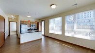 This single-floor space features an open-concept layout that seamlessly connects the kitchen and living area. The kitchen is outfitted with stainless steel appliances, including a full-size refrigerator, range, and microwave, complemented by warm wood cabinetry and a dark-topped peninsula that provides additional prep and dining space. Three distinctive red pendant lights hang above the peninsula, adding a bold accent against the neutral, warm-toned walls. A flush ceiling light fixture illuminates the living area, which benefits greatly from a large multi-pane window that invites abundant natural light and offers views of the surrounding urban environment. A secondary door adjacent to the kitchen provides additional access to the exterior. The hardwood-style flooring runs throughout the space, lending warmth and visual continuity between zones. The overall ambiance is clean, modern, and functional, with an efficient use of space that makes the open layout feel both inviting and practical for everyday living.