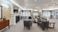 Located at 15 Princeton Dr in Hooksett, New Hampshire, this single-floor community clubhouse offers a thoughtfully designed gathering space with upscale finishes and a welcoming ambiance. The expansive main lounge is a standout feature, showcasing tray ceilings with ambient cove lighting, a vaulted ceiling section, and a classic white-mantled fireplace with a dark slate surround. A wall-mounted flat-screen TV anchors one side of the room, while generous carpeted seating areas are furnished with plush gray sofas, patterned armchairs, and tufted ottomans centered around a warm wood coffee table. Large windows with elegant drapery and a prominent arched window flood the space with natural light, offering views of the surrounding landscape. A casual dining nook and additional seating areas are seamlessly integrated into the lounge, creating versatile zones for socializing. The kitchen area is well-appointed with rich warm-toned wood cabinetry, granite countertops, mosaic tile backsplash, and stainless steel appliances. A kitchen bar with a dark quartz countertop and woven bar stools provides a stylish casual dining option, complete with pendant lighting above. Dark tile flooring in the kitchen contrasts beautifully with the lighter tones of the lounge, creating a cohesive yet distinct flow throughout the space.