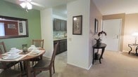 This single-floor residence offers a thoughtfully arranged layout that balances functionality and comfort. The dining area is carpeted and styled with warm, earthy tones, featuring a round wooden dining table with chairs — an inviting spot for casual meals. A soft green accent wall adds a touch of color and defines the space.

Adjacent to the dining area, the kitchen is accessible through a semi-open passageway, creating a connected yet distinct zone. The kitchen features dark wood cabinetry, a tile floor, a double sink with a window above, and a tasteful tile backsplash, providing a practical and classic cooking environment.

A central column and partial wall divide the dining space from a hallway area, which is finished with warm taupe-toned walls and continues with carpet flooring. A decorative console table adorned with a floral arrangement and framed artwork on the walls lend the hallway an elegant, curated ambiance.

Natural light filters through the space, complementing the neutral color palette of whites, greens, and taupes throughout. The overall design reflects a traditional residential aesthetic, combining functional areas with decorative touches that create a cohesive and livable environment.