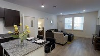 This single-floor residence is thoughtfully designed with an open-concept layout that integrates the kitchen, dining, and living spaces into one cohesive and functional area. The kitchen features a granite countertop with a breakfast bar, accommodating bar-height seating with sleek black chairs, making it an ideal spot for casual dining or entertaining. The countertop is complemented by clean white cabinetry and modern fixtures.

The living area is generously sized and anchored by a sectional sofa arrangement, providing ample seating. A decorative starburst mirror serves as a focal point on the white walls, adding a stylish accent to the space. An arc floor lamp enhances the ambient lighting alongside recessed ceiling lights that brighten the entire open area.

Light hardwood-style flooring runs throughout, lending warmth and continuity across the main living space. Natural light flows in through multiple windows fitted with white blinds, creating a bright and welcoming atmosphere. A doorway visible from the main area provides access to a bedroom, which features warm lighting and a wooden bed frame, suggesting a comfortable and private retreat.