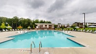 Located at 888 Horizon Boulevard in Athens, Georgia, this outdoor amenity space is centered around a generously sized resort-style swimming pool with crystal-clear blue water. The pool area is designed for both relaxation and recreation, featuring a separate shallow wading pool alongside the main pool, making it suitable for a wide range of users. The surrounding sun deck is finished in smooth, light-toned concrete and is outfitted with an extensive collection of sling-style lounge chairs and patio umbrellas in a mix of striped and solid orange patterns, providing ample shade and comfort throughout the day. The clubhouse building anchors one side of the pool area, constructed in a modern style with white siding, bold orange-framed floor-to-ceiling windows, and glass doors that offer views into the interior. Lush green lawns and mature trees border the perimeter, enclosed by black iron fencing that defines the space while maintaining open sightlines to the surrounding landscaping. Decorative plantings and young trees are interspersed throughout the grounds, adding a natural, park-like quality to the environment. The overall ambiance is clean, contemporary, and inviting, offering a well-appointed outdoor retreat within the broader community setting.