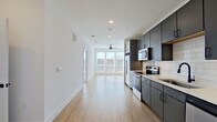 This thoughtfully designed one-bedroom, one-bathroom unit offers a clean, contemporary aesthetic across its efficiently organized single-floor layout. The open-concept living and kitchen area is anchored by light wood-style flooring and bright white walls, creating an airy, cohesive feel. A ceiling fan overhead and large windows at the far end of the living space flood the area with natural light while offering outdoor views.

The kitchen is well-appointed with dark gray cabinetry, light quartz countertops, a white subway tile backsplash, stainless steel appliances including a refrigerator, range, microwave, and dishwasher, and a matte black faucet over a single-basin sink. The contrast between the dark cabinetry and light surfaces lends a modern, polished character.

The bedroom features plush carpet flooring, a ceiling fan, and generous windows that bring in ample daylight. Additional areas visible in the floorplan include a dedicated hallway connecting the main living spaces to the bedroom and bathroom.

The bathroom is finished with large-format tile flooring, a walk-in glass-enclosed shower with marble-style tile surround and matte black hardware, a toilet, and a vanity with matching dark cabinetry and a white countertop.