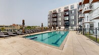 This outdoor amenity space centers around a generously sized lap pool with crisp, blue-tiled water and wide surrounding deck. The pool deck is finished in large-format concrete pavers, providing ample space for relaxation and movement around the perimeter. A substantial row of cushioned chaise lounges lines one side of the deck, offering dedicated sunbathing areas ideal for unwinding. Tall wooden post accents punctuate the deck, adding a distinctive design element that complements the contemporary architectural surroundings.

The space is enveloped by a modern multi-story residential building featuring a striking contrast of dark charcoal and light grey façades, large windows, and private balconies that overlook the pool area. A black metal fence borders the deck, providing safety while maintaining an open, airy feel. A separate gathering area is visible, complete with outdoor seating, a wall-mounted television, and string lighting, creating an ideal setting for social gatherings. Pool entry is made convenient via wide, tiled steps with stainless steel handrails. Panoramic city and treetop views extend beyond the building, enhancing the open-air atmosphere of this well-appointed outdoor retreat.