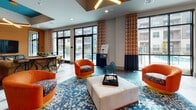 This single-floor community clubhouse is designed with a bold, contemporary aesthetic that blends vivid color palettes with thoughtful architectural detail. The main lounge area features plush navy blue wingback chairs arranged around a rustic wood coffee table, set atop a striking orange geometric rug. A large flat-screen TV is mounted against a richly textured dark accent wall, complemented by a vibrant mosaic art panel nearby. Floor-to-ceiling windows with patterned drapery flood the space with natural light and provide views of the surrounding exterior. A secondary seating area showcases rounded orange accent chairs, a central tufted ottoman, and a blue patterned area rug, creating a relaxed gathering spot. The gaming and dining zone features a multi-toned stacked-stone accent wall with an embedded flat-screen TV, a round table with upholstered blue chairs, and a pool table in the background. Geometric pendant and recessed lighting with gold-accented ceiling details add visual interest throughout. A kitchenette area is equipped with light wood cabinetry, a quartz countertop, and appliances, finished with an intricate geometric tile backsplash. Large-format tile flooring unifies the entire space, delivering a polished, cohesive feel throughout this well-appointed communal facility.