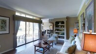 This single-floor residence offers an open-concept layout that integrates the living, dining, and sitting areas into one cohesive space. The living room is anchored by a comfortable sectional sofa and a classic wooden coffee table, positioned atop a neutral area rug that defines the seating zone. A standout architectural feature is the built-in arched shelving unit along one wall, providing both display and storage space with decorative items and books neatly arranged across its shelves.

Large sliding glass doors flood the interior with natural light and open onto a balcony, offering views of lush greenery beyond. Curtain panels frame the doors, adding a layered, finished look to the space. The warm taupe wall tones complement the wood flooring and neutral furnishings, creating a cohesive and inviting ambiance throughout.

Adjacent to the living area, a dining space accommodates a dark wood table and matching chairs, illuminated by a pendant light above. Crown molding runs along the ceiling perimeter, adding a refined architectural detail that ties the overall design together elegantly.