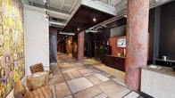 This single-floor commercial space makes a striking first impression with its industrial-chic design aesthetic. The main corridor stretches generously, anchored by large-format stone-look tile flooring that lends a sense of scale and openness. Overhead, an exposed ceiling reveals ductwork, piping, and structural elements painted in a neutral gray, complemented by a suspended dark wood slat feature ceiling that adds warmth and visual definition to the reception area.

Bold, deep red accent walls and richly textured columns with a raw, distressed finish create a dramatic contrast against the lighter flooring and white walls, giving the space a strong and memorable character. A built-in dark wood reception counter is positioned prominently, accompanied by wall-mounted flat-screen displays and tasteful greenery that soften the industrial elements.

Track lighting and pendant spotlights are strategically placed throughout, ensuring the space is well-lit while maintaining its atmospheric quality. A seating area near the entrance features woven accent chairs alongside a small side table, offering a welcoming spot for visitors. The overall ambiance balances rugged, warehouse-inspired design with polished commercial functionality, resulting in a space that feels both dynamic and professionally curated.