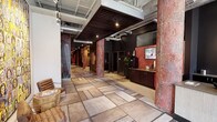 This single-floor commercial space makes a striking first impression with its industrial-chic design aesthetic. The main corridor stretches generously, anchored by large-format stone-look tile flooring that lends a sense of scale and openness. Overhead, an exposed ceiling reveals ductwork, piping, and structural elements painted in a neutral gray, complemented by a suspended dark wood slat feature ceiling that adds warmth and visual definition to the reception area.

Bold, deep red accent walls and richly textured columns with a raw, distressed finish create a dramatic contrast against the lighter flooring and white walls, giving the space a strong and memorable character. A built-in dark wood reception counter is positioned prominently, accompanied by wall-mounted flat-screen displays and tasteful greenery that soften the industrial elements.

Track lighting and pendant spotlights are strategically placed throughout, ensuring the space is well-lit while maintaining its atmospheric quality. A seating area near the entrance features woven accent chairs alongside a small side table, offering a welcoming spot for visitors. The overall ambiance balances rugged, warehouse-inspired design with polished commercial functionality, resulting in a space that feels both dynamic and professionally curated.