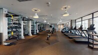 This expansive fitness center is thoughtfully designed to accommodate a wide range of workout disciplines within a single, open floor plan. The space is anchored by large floor-to-ceiling windows along one wall, flooding the interior with natural light and offering views of the surrounding cityscape. Rubber flooring throughout provides both durability and comfort underfoot across all zones.

The cardio section lines the window wall and features an impressive array of treadmills, elliptical trainers, stationary bikes, and a stair climber, all oriented to take advantage of the outdoor views. Pendant lighting fixtures hang from the white ceiling, complementing the recessed lighting and contributing to a bright, modern atmosphere.

The strength training area is well-stocked with a comprehensive dumbbell rack, a cable and multi-functional rig system, and weight benches positioned in front of floor-to-ceiling mirrors that amplify the sense of space. A dedicated functional training zone includes TRX suspension systems, medicine balls, and open floor space for dynamic movement.

A separate studio room, enclosed by glass-panel walls, features a ballet barre, mirrored walls, and a wall-mounted television, making it suitable for group classes or individual training. A wall-mounted television, water station, and restroom access round out the practical amenities of this polished, professionally appointed facility.