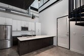 Located at 1143 First Ave S in Birmingham, Alabama, this two-story space blends industrial character with modern finishes throughout. The ground floor showcases soaring ceilings with exposed concrete beams and ductwork, polished concrete floors, and crisp white walls that create an open, loft-like atmosphere. A well-appointed kitchen anchors the main level, featuring a large island with a white quartz countertop and integrated stainless steel sink, dark walnut lower cabinetry contrasted by white upper cabinets, subway tile backsplash, and stainless steel appliances including a refrigerator, range, and built-in microwave. Distinctive pendant lighting adds a contemporary accent above the island. A full bathroom on this level includes a glass-enclosed walk-in shower with white subway tile, a white vanity with ample storage, and polished concrete flooring. A striking open staircase connects the two floors, leading to the upper level where polished concrete floors continue beneath a ceiling fan. The second floor is bathed in natural light from a bank of large windows offering views of the surrounding neighborhood. Black metal railings lining the open stairwell provide an industrial-modern design element that unifies both levels cohesively.