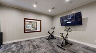 This fitness facility is thoughtfully designed to support a wide range of workout routines within a single, cohesive space. The interior is finished with neutral gray walls, recessed lighting, and durable patterned carpet flooring throughout, creating a clean and focused environment for exercise.

The spin studio area is outfitted with multiple stationary cycling bikes and a large wall-mounted flat-screen television, ideal for guided cycling sessions and virtual workouts. A large framed window connects this area to the main fitness floor, providing visual openness between the two zones.

The main gym area features a Life Fitness cable and strength machine, along with a comprehensive functional training rack stocked with a full selection of medicine balls, stability balls, dumbbells, and kettlebells, catering to both strength and conditioning workouts.

Entry to the facility is through elegant wood-framed glass doors, which lead into a transitional corridor connecting to an adjacent space accented with distinctive green patterned wallcovering and a small reception counter. The overall design balances functionality with a polished aesthetic, making this fitness center a well-appointed and inviting space for daily training.