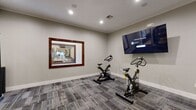 This fitness facility is thoughtfully designed to support a wide range of workout routines within a single, cohesive space. The interior is finished with neutral gray walls, recessed lighting, and durable patterned carpet flooring throughout, creating a clean and focused environment for exercise.

The spin studio area is outfitted with multiple stationary cycling bikes and a large wall-mounted flat-screen television, ideal for guided cycling sessions and virtual workouts. A large framed window connects this area to the main fitness floor, providing visual openness between the two zones.

The main gym area features a Life Fitness cable and strength machine, along with a comprehensive functional training rack stocked with a full selection of medicine balls, stability balls, dumbbells, and kettlebells, catering to both strength and conditioning workouts.

Entry to the facility is through elegant wood-framed glass doors, which lead into a transitional corridor connecting to an adjacent space accented with distinctive green patterned wallcovering and a small reception counter. The overall design balances functionality with a polished aesthetic, making this fitness center a well-appointed and inviting space for daily training.