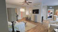 This single-floor space offers a thoughtfully designed open-concept layout that maximizes functionality and comfort. The living area is anchored by rich hardwood-style flooring that extends throughout, lending warmth and a cohesive aesthetic to the entire space. A ceiling fan with a sleek finish provides both style and practicality overhead, while a bold arc floor lamp adds an elegant lighting accent to the living zone. A plush sofa and woven area rug create a welcoming and relaxed seating arrangement.

Transitioning seamlessly from the living area, a compact bar-height counter with stools serves as a natural dividing element, ideal for casual dining or entertaining. Pendant lights hang above this counter, adding a decorative touch and targeted illumination. A wall-mounted flat-screen television is positioned prominently, making it easily viewable from both the seating and dining areas.

The space also features a wall-mounted mini-split HVAC unit for efficient climate control, and multiple doorways lead to additional rooms, suggesting a well-connected and practical floor plan. Neutral wall tones throughout keep the atmosphere bright and versatile, making this a clean, modern, and highly livable space.