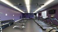 Located at 3800 Bostic Dr in Greenville, NC, this fitness center offers a well-equipped, single-floor workout facility designed to support a full range of exercise routines. The main workout area is generously sized, providing ample space for both cardio and strength training activities. The room is finished with durable dark rubber flooring, ideal for high-traffic gym use, and the walls are painted in a bold, energizing deep purple that gives the space a distinctive and motivating atmosphere. A row of treadmills lines one side of the floor, offering dedicated space for cardiovascular workouts, while free weights, benches, and additional cardio machines are arranged throughout the room to accommodate a variety of fitness needs. Overhead, a series of ceiling fans are evenly distributed across the ceiling alongside bright panel lighting, ensuring consistent airflow and visibility throughout the space. A wall-mounted television is positioned for easy viewing during workouts. Natural light enters through windows along one wall, adding to the openness of the environment. The overall layout is clean, organized, and functional, making this fitness center a practical and well-appointed destination for exercise.
