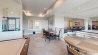 Located at 301 S Calhoun Pl in Star, Idaho, this single-floor community space offers a thoughtfully designed layout encompassing a game room, library, and connecting hallway. The interior features a clean, contemporary aesthetic with neutral gray and taupe wall tones, plush carpeted flooring, and recessed lighting throughout, creating a warm and welcoming atmosphere.

The game room serves as a social hub, furnished with round tables and cushioned chairs ideal for casual gatherings, alongside a shuffleboard table and open areas for activity. A sleek metal railing lines the open-concept edges, adding a modern architectural touch while maintaining visual connectivity across the space.

The library area provides a quieter retreat, furnished with a leather sofa, framed artwork, and tasteful greenery that lend a relaxed, comfortable feel. An adjacent dining area features a solid wood table beneath a contemporary black chandelier, bathed in natural light streaming through large black-framed windows that frame views of the surrounding Star, Idaho landscape.

Throughout the space, tray ceilings and elevated sightlines contribute to a sense of openness and volume. The interplay of natural light, modern furnishings, and cohesive neutral tones creates a versatile environment well-suited for both collaborative and quiet activities. The overall ambiance is polished yet approachable, reflecting a modern community-centered design.
