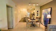This single-floor residence showcases an open-concept design that seamlessly connects the dining area and kitchen into one cohesive living space. The dining area is anchored by a round pedestal table surrounded by upholstered chairs in a soft grey finish, creating an inviting and refined atmosphere. Decorative wall accents, including circular ornamental pieces and framed artwork, add character to the sage-toned accent wall nearby.

The kitchen is efficiently designed and features stainless steel appliances, including a full-size refrigerator and microwave, complemented by light-toned cabinetry. A kitchen island with bar stool seating provides additional prep space and casual dining options. Track lighting runs along the ceiling, delivering focused illumination throughout both the kitchen and dining zones.

The flooring transitions from plush, neutral-toned carpet in the dining area to hard flooring in the kitchen, defining each functional zone while maintaining visual continuity. Crown molding along the ceiling adds a polished architectural detail, enhancing the overall sense of quality. A hallway visible from the main space leads to additional rooms, suggesting a well-organized and practical floor plan throughout.