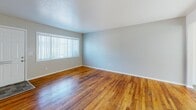 This single-floor home offers a clean, functional layout with consistent design elements carried throughout. Rich, warm-toned hardwood flooring runs across the main living areas and bedrooms, adding character and a sense of continuity to the space. The living area is open and inviting, featuring light gray walls, white baseboards, a textured ceiling, and a sliding glass door that floods the room with natural light and provides views of the surrounding greenery outside. A floor vent with a built-in register points to a central HVAC system serving the home's climate control needs. The bedrooms are well-proportioned, each featuring the same hardwood flooring and wide windows fitted with vertical blinds that allow for flexible light control and privacy. The kitchen is equipped with white cabinetry, gray countertops, a stainless steel electric range with an overhead exhaust hood, and a refrigerator — providing a practical and tidy cooking environment. The bathroom includes a full tub-shower combination with large-format beige wall tile, a toilet, a single-sink vanity with dark countertop and white cabinetry, and a small window for natural ventilation. The overall ambiance of the home is bright, clean, and move-in ready, with a straightforward layout that makes efficient use of space.