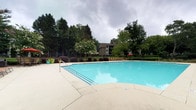 This outdoor amenity space centers around a generously sized swimming pool with clear, blue water and a smooth concrete surrounding deck. Entry into the pool is made easy via built-in stair steps and a sturdy handrail, accommodating swimmers of varying comfort levels. The deck provides ample space for relaxation and is lined with lounge chairs and an umbrella, offering residents a comfortable place to unwind poolside. The area is thoughtfully landscaped with a variety of mature trees, flowering shrubs, and decorative potted plants that add color and a sense of privacy to the environment. A low, dark metal fence encloses the pool area, providing both safety and definition to the space. The surrounding greenery creates a natural, park-like backdrop, with tall trees framing the space and contributing to a serene, shaded atmosphere. A multi-story residential building is visible in the background, situating this pool area as a shared community amenity. The overall ambiance is open, refreshing, and welcoming, making it an ideal outdoor retreat for relaxation and recreation during warmer months.