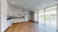 This single-floor unit is designed with a clean, modern aesthetic that emphasizes open living. The kitchen and living area share an open-plan layout, creating a spacious and connected environment. The kitchen is well-appointed with white cabinetry extending to the ceiling for ample storage, dark countertops, a stainless sink, a dishwasher, a white refrigerator, and a white range with an overhead microwave. Recessed lighting and a ceiling fan provide both illumination and air circulation throughout the main living space. Warm-toned wood-look flooring runs consistently through the kitchen and living areas, contributing to a cohesive, welcoming atmosphere. Large sliding glass doors fitted with vertical blinds open to a balcony, allowing abundant natural light and outdoor views into the main living space. A separate bedroom features plush carpeting, a ceiling fan, and a large window with vertical blinds that admit generous natural light. The full bathroom is finished with light marble-style tile flooring, a white vanity with under-sink storage, a large mirror, and a shower-tub combination. Additional built-in storage cabinetry is present within the unit, adding to its practical functionality.