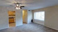 This single-floor residence offers a practical and comfortable layout designed for everyday living. The main living area is finished with plush wall-to-wall carpeting that adds warmth underfoot, while a ceiling fan with integrated light fixture provides both ventilation and ambient illumination. A wide window dressed with horizontal blinds allows natural light to fill the room, creating a bright and airy atmosphere. A wall-mounted heating unit beneath the window ensures year-round comfort. The living space flows naturally toward an entry door, providing a welcoming transition point. Adjacent to the living area, the kitchen is equipped with wooden cabinetry, a gas range, and a built-in microwave above the stove, offering essential functionality in a compact footprint. The neutral, light-toned walls throughout the space lend a clean and versatile backdrop, making it easy to personalize. The overall design is straightforward and unpretentious, prioritizing utility and livability in a well-maintained single-level floor plan.