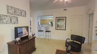 This single-floor residence offers a well-designed open layout that maximizes both functionality and comfort. The living area immediately draws attention with its soaring vaulted ceilings, which lend a sense of spaciousness and airiness to the room. A ceiling fan is centrally positioned overhead, providing both practical ventilation and a casual aesthetic touch.

The living space features plush wall-to-wall carpeting and is furnished with a classic wooden media console, decorative accents, and wall art that adds personality to the neutral, light-toned walls. A pass-through window connects the living area to the kitchen, where warm wood-toned cabinetry and a raised breakfast bar with two elegant wrought-iron bar stools create an inviting casual dining spot. This architectural detail enhances the open-concept feel while maintaining a subtle distinction between spaces.

The kitchen, visible through the pass-through, is compact yet functional, with ample cabinetry and counter space. Natural light filters through the windows, brightening the interior and complementing the clean white walls throughout. A standard interior door provides access to additional areas of the home. Overall, the space presents a comfortable, well-lit, and thoughtfully arranged single-floor layout suited for everyday living.